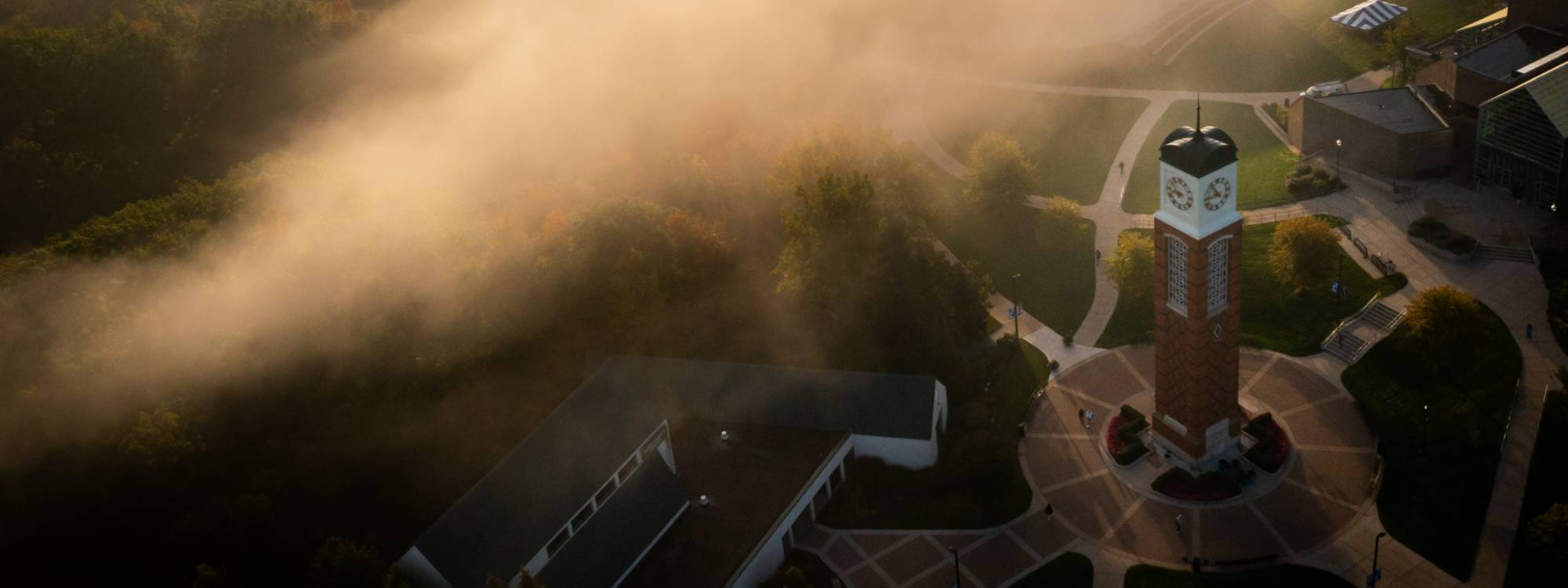 An aerial photo of the Allendale campus shows the clock tower and Cook DeWitt Center as fog rises above the ravines.